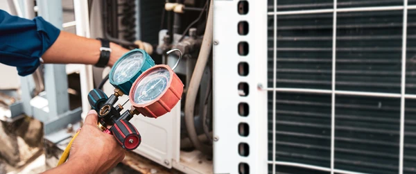 Technicien en combinaison bleue utilisant une pince ampèremétrique pour effectuer un diagnostic électrique sur le panneau de contrôle ouvert d'une unité extérieure de climatisation split-system blanche fixée au mur, avec câblages visibles et connecteurs internes révélés, illustrant une intervention de maintenance sur équipement climatique quel que soit le fabricant.