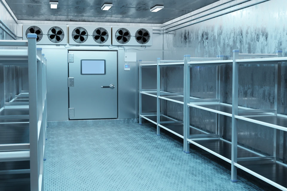 Technicien frigoriste en combinaison bleue étudiant les plans d'installation d'une chambre froide, debout dans un local commercial vide montrant les dimensions au sol, les points d'alimentation électrique et les contraintes architecturales du plafond et des parois. Il tient un dossier technique ou son téléphone, pointant une zone du local. Vue générale du local avec au second plan les équipements d'accès et les portes. Éclairage naturel. Aucun personne autre que le technicien.